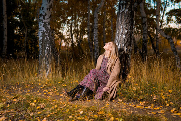 Young beautiful smiling woman in long coat sitting on the ground in autumn forest.