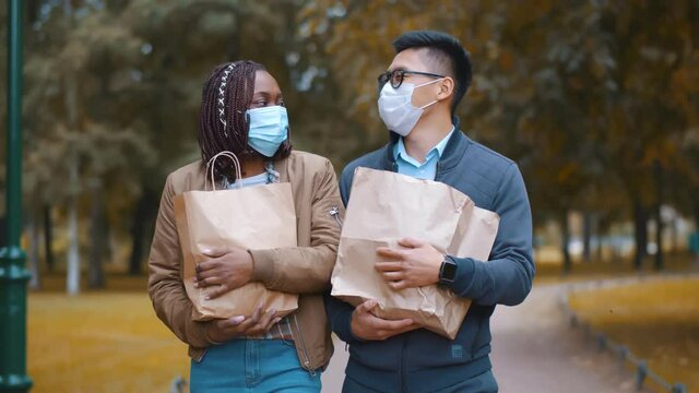 Happy multiethnic young couple in safety mask with groceries walking in park
