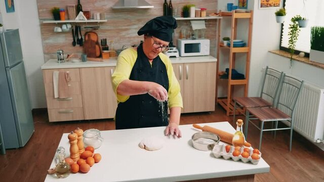 Retired housewife with bonete and kitchen apron adding sifted flour on the dough. Elderly senior baker with uniform sprinkling, sifting, spreading ingredients by hand baking homemade pizza and bread.