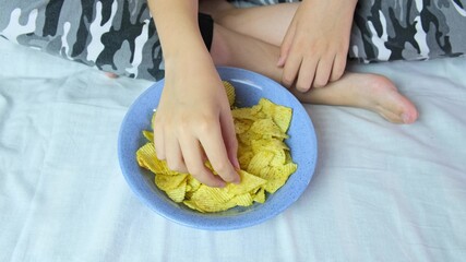 A boy sitting on a sofa at home and eating crispy potato chips from a bowl, close up view