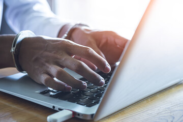 Close up hands of hacker with handcuffs above notebook keyboard, punishment for cybercrime,...