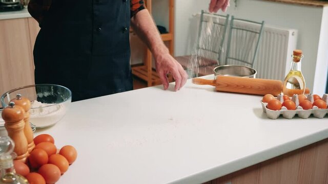 Elderly man with bonete and kitchen apron sieving flour on table ready for cooking. Retired senior baker with uniform sprinkling, sifting, spreading rew ingredients baking homemade pizza and bread.
