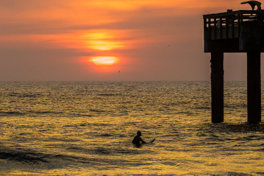 Sunrise over the atlantic ocean at St Augustine Beach in Florida, a fishing pier on the right.  A surfer waits for a big wave.