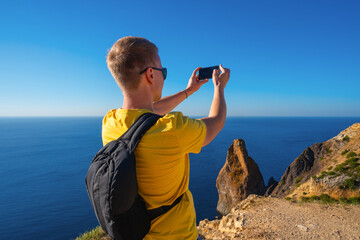 Fototapeta na wymiar A young man with a backpack takes photos with a smartphone of the view of the rocks and the blue sea