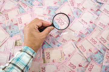 Ukrainian banknotes are checked under a magnifying glass. conceptual photo. Female hand with magnifying glass. 200 hryvnia background.
