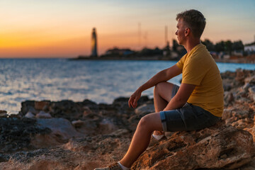 Fototapeta na wymiar A young man sits thoughtfully on a rock and looks at the sunset, with a lighthouse by the sea in the background