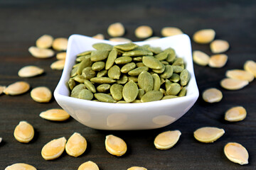 Closeup a bowl of tasty and healthy roasted pumpkin seeds with raw seeds scattered on wooden table