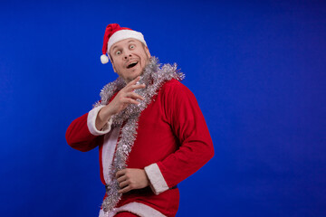 Male actor in a suit and hat of Santa Claus with a silver tinsel garland dancing and posing on a...