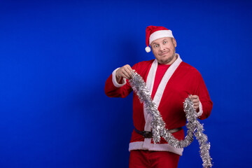 Male actor in a suit and hat of Santa Claus with a silver tinsel garland dancing and posing on a...