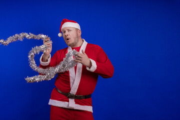 Male actor in a suit and hat of Santa Claus with a silver tinsel garland dancing and posing on a...