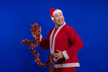 Male actor in a suit and hat of Santa Claus with a red tinsel garland dancing and posing on a blue...