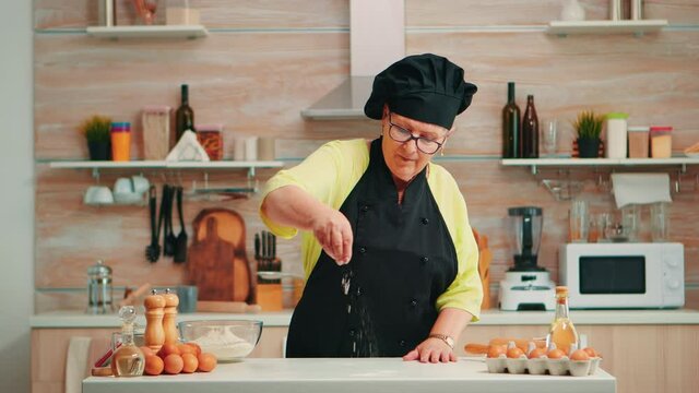 Woman is sifting flour on wooden table in modern kitchen. Happy elderly baker with bonete preparing raw ingredients to baking homemade cake sprinkling, sieving wheat flour by hand