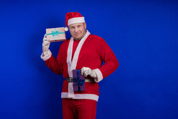 Emotional male actor in a costume of Santa Claus holds gift boxes in his hands and poses on a blue...