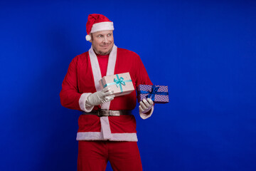 Emotional male actor in a costume of Santa Claus holds gift boxes in his hands and poses on a blue...