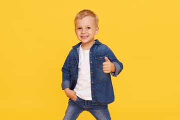 Joyful little child boy in denim clothes shows thumb up, OK symbol on yellow background. Advertising of childrens products and happy childhood