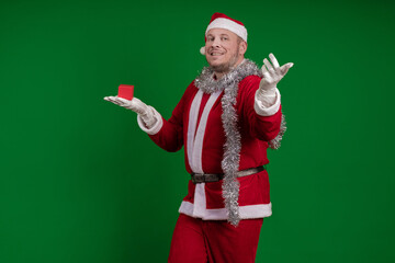 Emotional male actor in a costume of Santa Claus holds a small red gift box in his hands and poses...