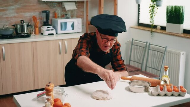 Professional old chef sprinkling flour on dough in home kitchen. Elderly senior baker with bonete and uniform sieving, sifting, spreading rew ingredients by hand, baking homemade pizza and bread.