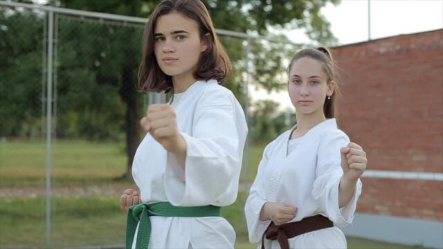 Two young girls stand in classic karate poses on the sports ground and look at the camera. Close-up. Slow motion. The camera moves from side to side and changes the shooting angle