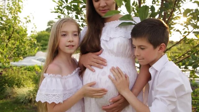 A pregnant mother with two children are walking in the garden near the house. Family with pregnant mom in the summer in nature. Children hugging pregnant belly