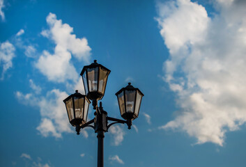 Fototapeta na wymiar Old vintage black decorative lantern with clear glass on pillar. Three street lamps on one pole. Sunny blue sky with white clouds