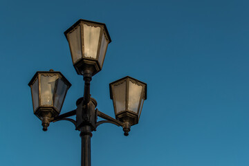 Fototapeta na wymiar Old vintage black decorative lantern with clear glass on pillar. Three street lamps on one pole in sunset light. Blue sky