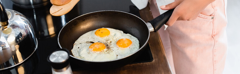 cropped view of woman holding kitchen tongs near eggs on frying pan, panoramic shot
