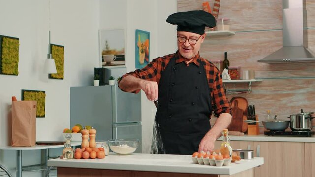 Experienced baker spreading flour in home kitchen for food preparation. Retired elderly chef with bonete and apron sprinkling, sieving sifting raw ingredients by hand baking homemade pizza, bread.