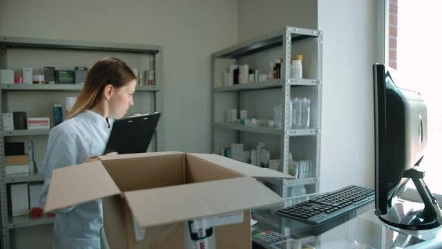Attractive caucasian woman unpacking delivery with medicine in pharmacy. Female with text tablet checking stuff in carton box and rowing medicines on showcase shelfs.