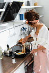 curly woman holding kitchen tongs while cooking eggs on frying pan in kitchen