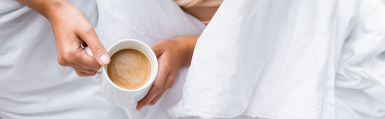 cropped view of woman holding cup with coffee in bed, panoramic shot