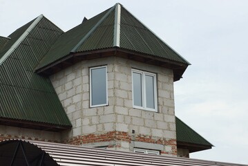 attic of a gray private brick house with windows under a green tiled roof against the sky