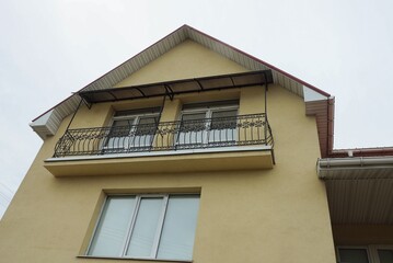 one open black iron balcony on the brown wall of a private house with white windows against a gray sky