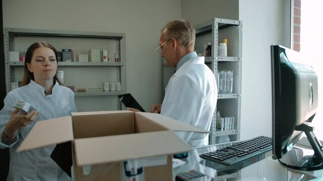 Two pharmacists collecting medicine items into delivery box for online customer. Young woman and mature man in white robes working as employees indoor of modern pharmacy interior.