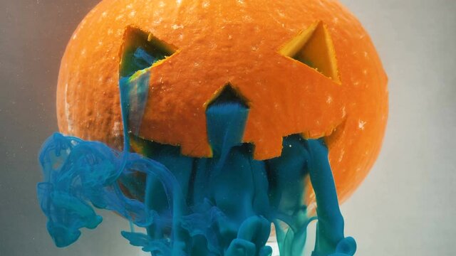 Scary Halloween orange pumpkin decoration with dense blue paint poured from cut mouth and nose holes in water extreme closeup