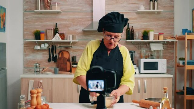 Woman communicating with subscribers through video camera while kneading dough. Retired blogger chef influencer using internet technology shooting blogging on social media with digital equipment