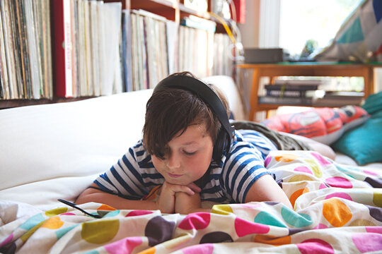 A young boy lies on bed with headphones using a tablet device