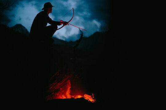 one adult male archer sits by the fire with his bow and arrow at dusk in the forest