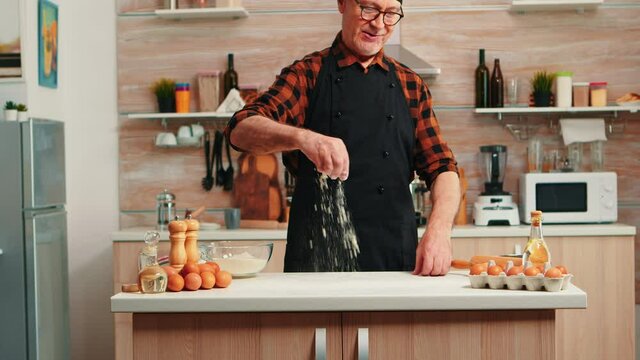 Elderly baker flouring table to bake and make delicious cookies. Retired senior chef with bonete and apron in uniform sprinkling sieving sifting raw ingredients by hand baking homemade pizza, bread