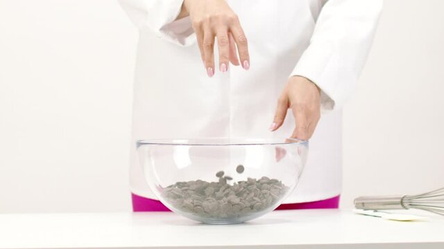 female chef pours handful chocolate couverture callets in glass bowl slow mo on white. flying Chocolate drops for preparation premium handmade candies, truffles, chocolates and sweets close up