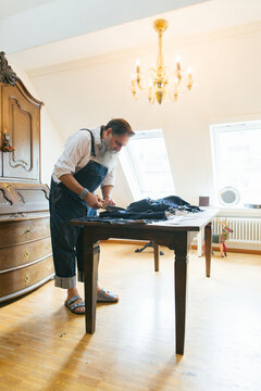 Indoor Portrait of Cool Senior Man Cutting Denim Fabric on Living Room Table