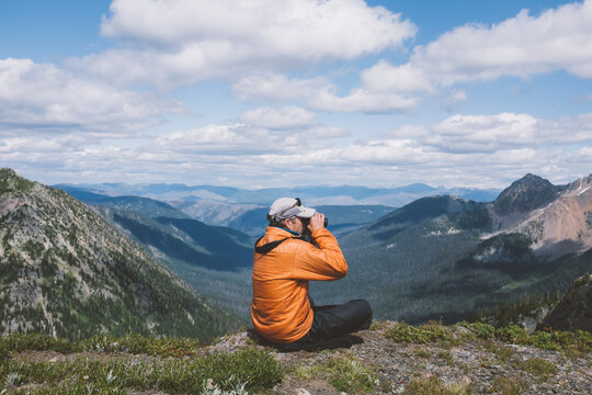 Man sitting on ledge, looking through binoculars at vast mountain range
