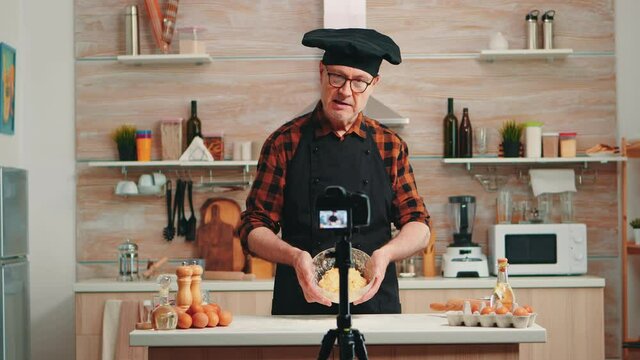 Mature experienced baker explaining recipe for audience recording tutorial for social media. Retired blogger chef influencer using internet technology communicating, shooting with digital equipment