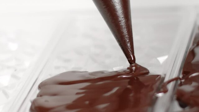 Close-up pouring dark chocolate liquid into transparent plastic mould. Filling out plastic bar-shaped designed mold with premium melted chocolate. Homemade organic chocolate production, slow motion