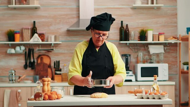 Woman baker using flour metallic sieve preparing homemade cakes. Happy elderly chef with bonete preparing raw ingredients to baking traditional bread sprinkling, sieving in the kitchen.