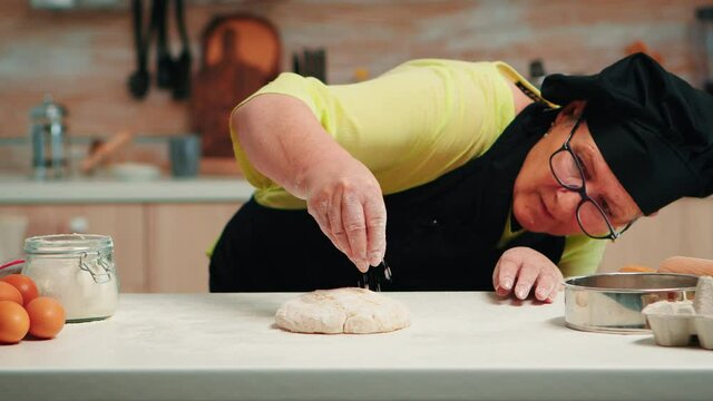 Woman with bonete and kitchen apron is occupied with dough preparation. Retired senior baker with apron, kitchen uniform sprinkling, sifting, spreading flour with hand baking homemade pizza and bread.