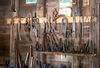 A collection of vintage blacksmith tools hung on racks on a wooden wall in a stone barn. The steel...