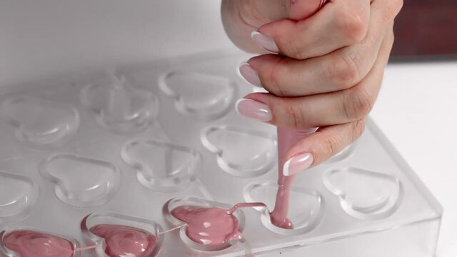 Closeup chocolatier pouring ruby melted chocolate into confectionery mold with pastry bag, making exclusive handmade chocolate candies. Luxury pink hand-crafted heart shaped candies. Slow motion
