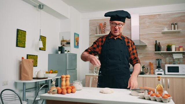 Grandfather sprinkling on the table sifted flour in modern kitchen. Elderly senior baker with bonete and uniform sieving, sifting, spreading rew ingredients on dough, baking homemade pizza and bread.