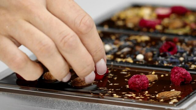 Close-up chocolatier decorating of luxury handmade chocolate bars with spices, dry berries and nuts. Making delicious dark organic hand-crafted chocolate, pastry shop, confectionery, slow motion
