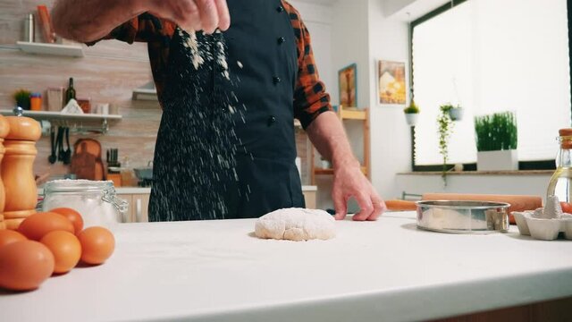 Close up of man sieving flour on bread dough on wooden table in the kitchen. Elderly senior baker with bonete and uniform sprinkling, sifting, spreading rew ingredients on dough, baking homemade pizza